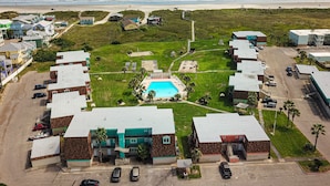 An aerial view of a coastal residential area with a swimming pool, multiple buildings, and a beach in the background. - Walk to Sandfest, Shared Pools, Boardwalk to Beach (Port Aransas)