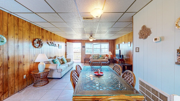 A spacious living room with a large glass-top dining table, wooden paneling on the walls, and a ceiling fan. - Walk to Sandfest, Shared Pools, Boardwalk to Beach (Port Aransas)