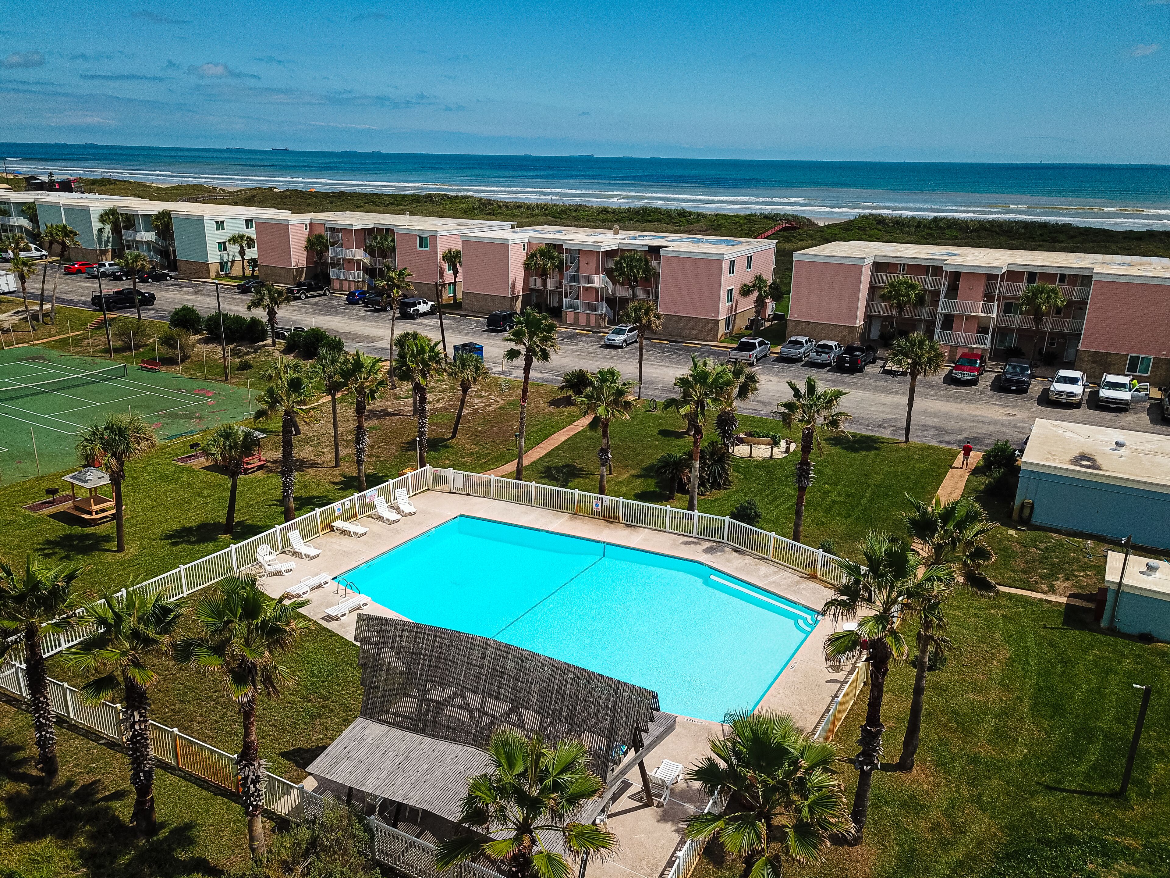 A pool area with a clear view of the ocean, surrounded by palm trees and a tennis court.