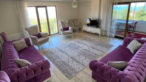 A spacious living room with a large sofa set, a television, and a view of the outdoors. - Menekse Apart 2 (Trabzon)