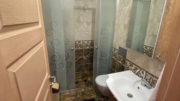 Un baño con un lavamanos blanco, un inodoro y una ducha con puerta de vidrio. Hay una alfombra roja en el suelo y una puerta de madera.