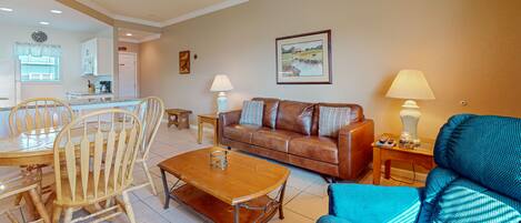 A living room with a brown leather sofa, a blue armchair, a wooden coffee table, and a dining area with a wooden table and chairs.