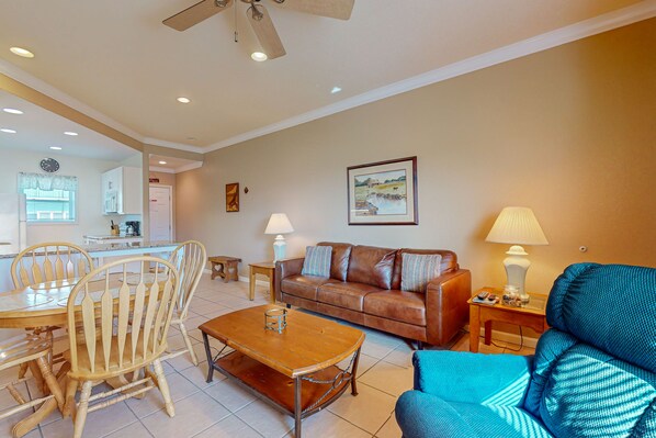 A living room with a brown leather sofa, a blue armchair, a wooden coffee table, and a dining area with a wooden table and chairs. - Walk to Sandfest, Shared Pools, Boardwalk to Beach (Port Aransas)