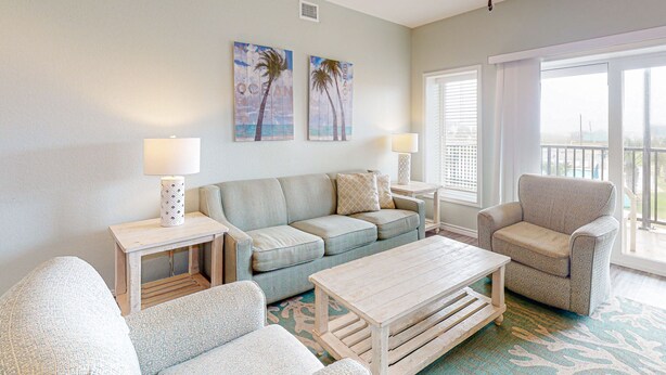 A living room with a sofa, armchairs, a coffee table, and two side tables with lamps. There are two wall-mounted paintings of palm trees and the word "OCEAN". A ceiling fan is visible, and the room has a large window with blinds. - Walk to Sandfest, Shared Pools, Board Walk to Beach (Port Aransas)