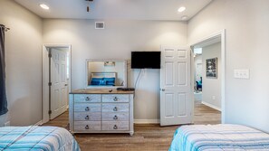 Quarto com duas camas, uma cômoda, TV fixada na parede e porta para outro espaço.