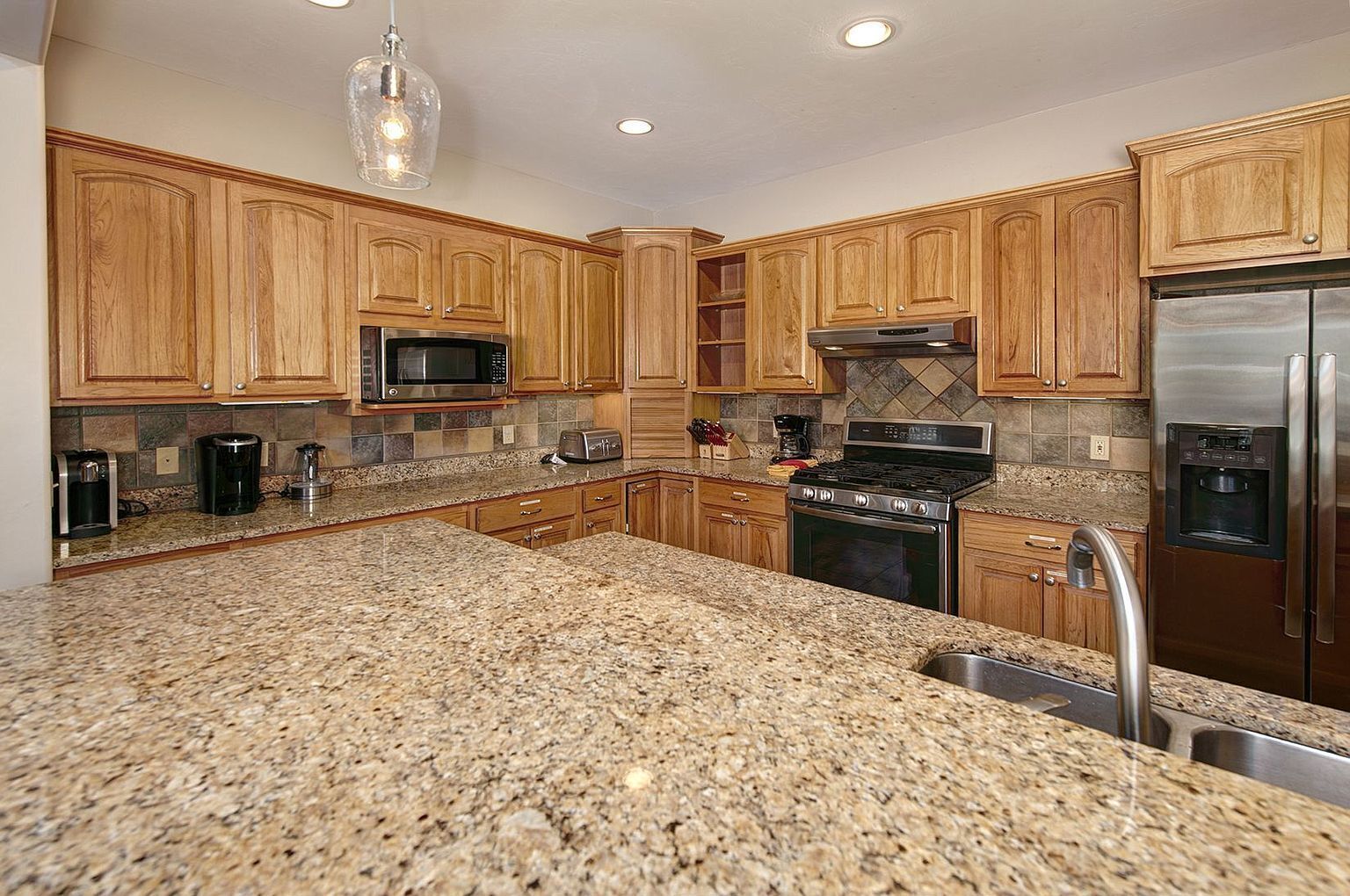 A kitchen with granite countertops, wooden cabinets, stainless steel appliances, and a hanging light fixture.