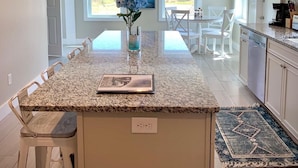 A modern kitchen with a granite island, pendant lighting, and a view of the outdoors through large windows. - Let the Sea Set You Free (South Kingstown)