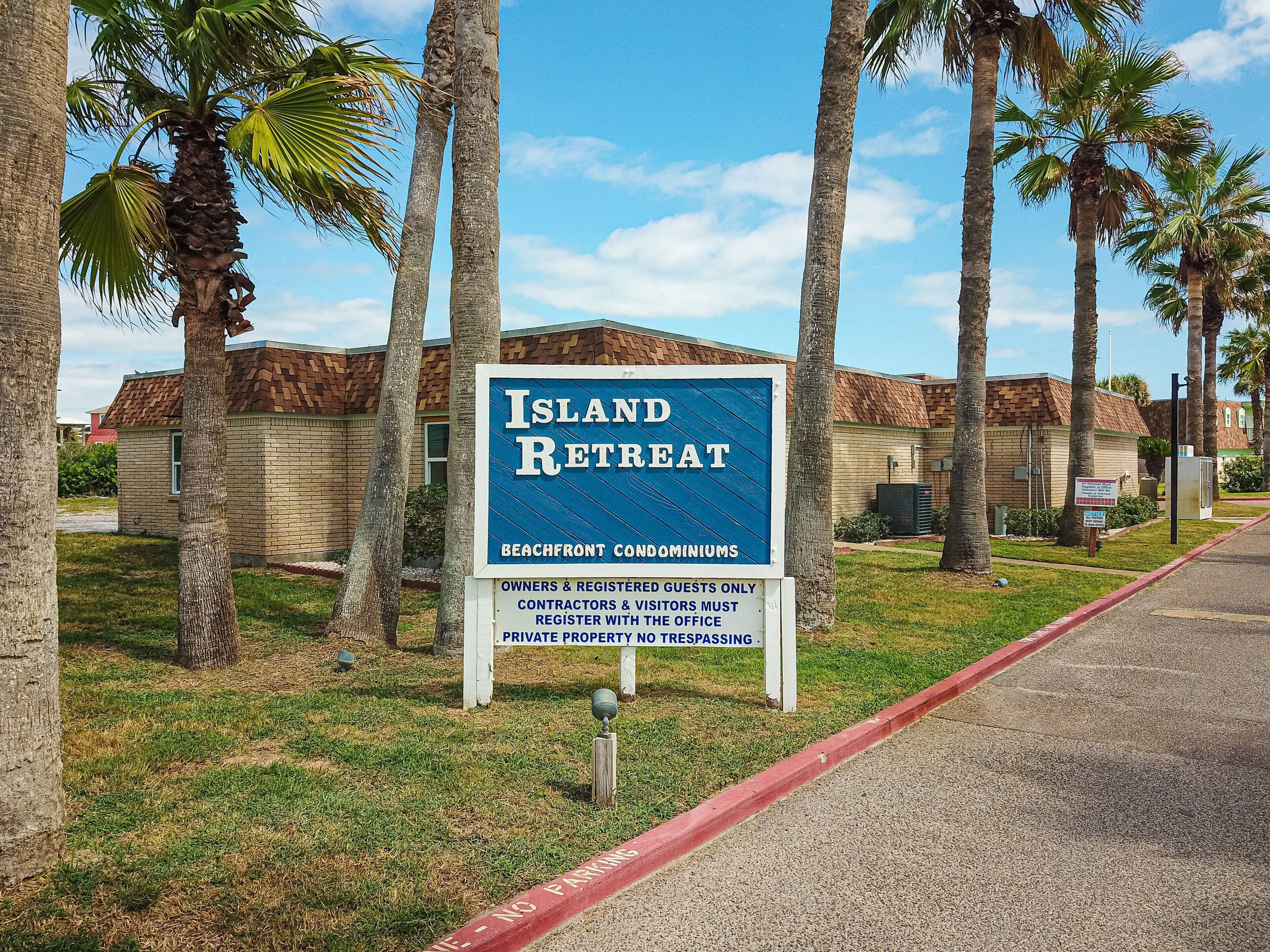 Une enseigne indiquant « Island Retreat », un complexe de condominiums situé en bord de mer, avec des palmiers et un ciel dégagé.