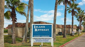 A sign for Island Retreat, a beachfront condominium complex, with palm trees and a clear sky. - Walk to Sandfest, Shared Pools, Boardwalk to the Beach (Port Aransas)