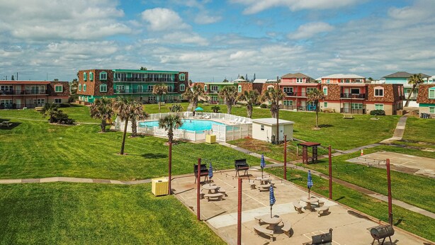A pool area with lounge chairs and umbrellas, surrounded by a grassy lawn and a building complex in the background. - Walk to Sandfest, Shared Pools, Boardwalk to the Beach (Port Aransas)