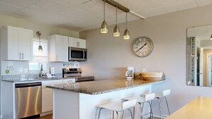 A modern kitchen with white cabinets, stainless steel appliances, a clock, and a bar area with stools. - Walk to Sandfest, Shared Pool, Boardwalk to the Beach (Port Aransas)