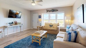 A living room with a ceiling fan, a television, a sofa, a coffee table, and a window with a view. - Walk to Sandfest, Shared Pool, Boardwalk to the Beach (Port Aransas)