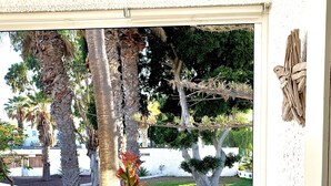A glass table with chairs on a balcony overlooking a garden with palm trees and a white fence.