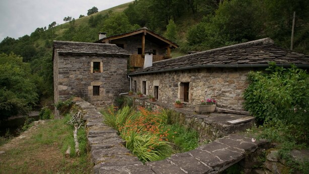 A rustic stone building with a wooden roof, a small balcony with potted plants, and a stone pathway leading to a water channel. - Molino de Yera (Vega de Pas)