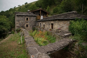Un edificio rústico de piedra con tejado de madera, un pequeño balcón con plantas en macetas y un camino de piedra que conduce a un canal de agua.