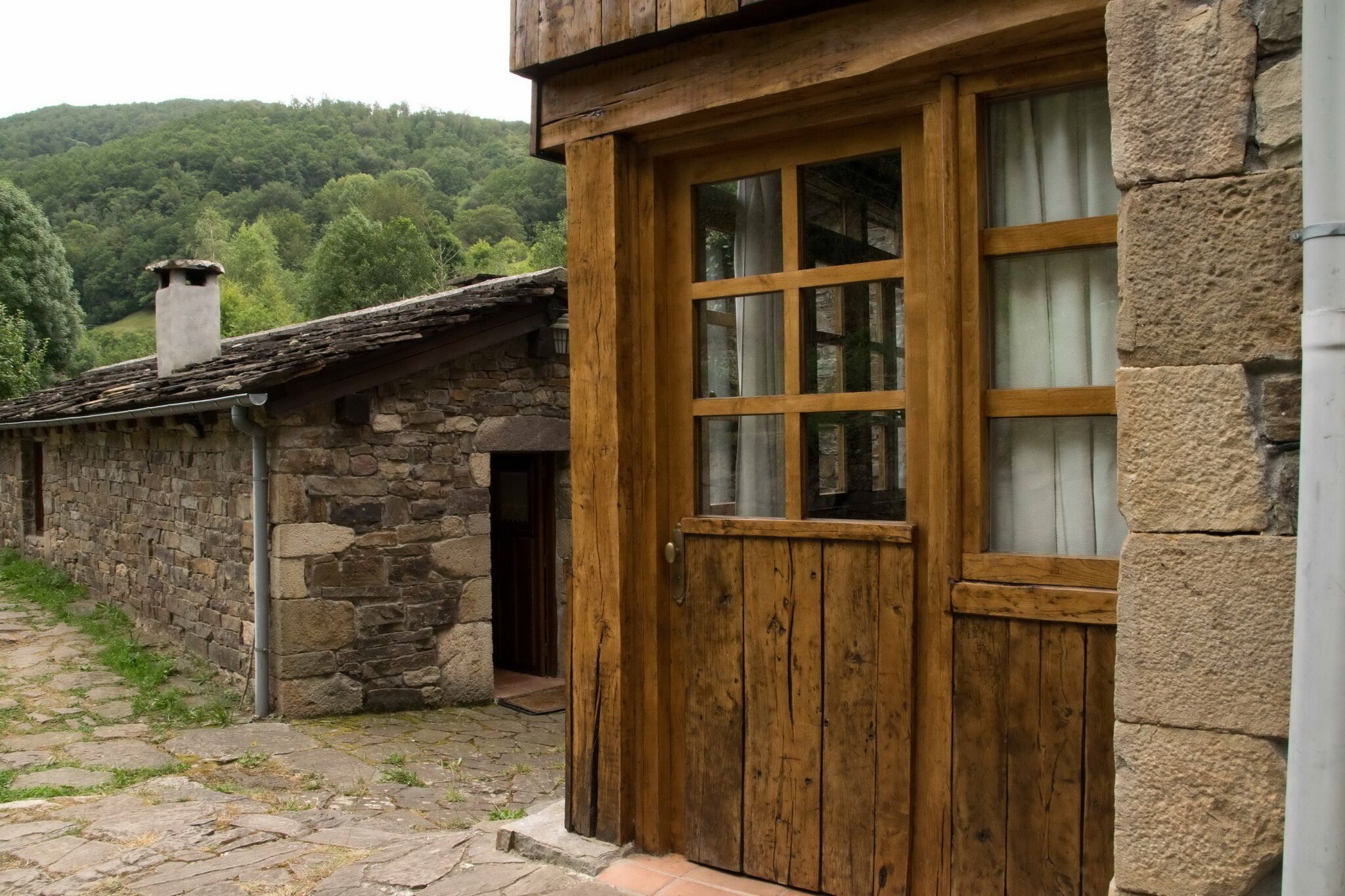 A rustic stone building with a wooden door and window, set in a rural area with a stone pathway and greenery in the background.