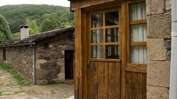 A rustic stone building with a wooden door and window, set in a rural area with a stone pathway and greenery in the background.