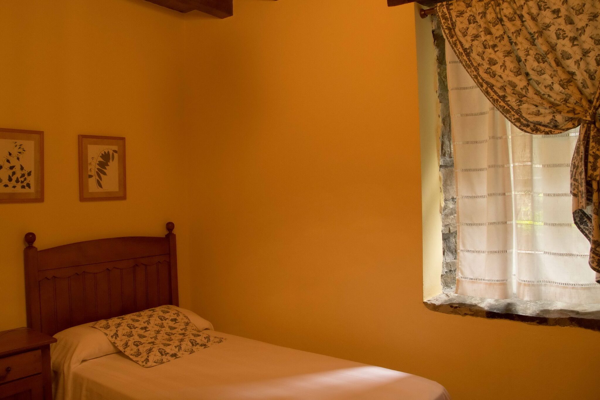 A bedroom with a wooden bed, a nightstand, and a window with patterned curtains.