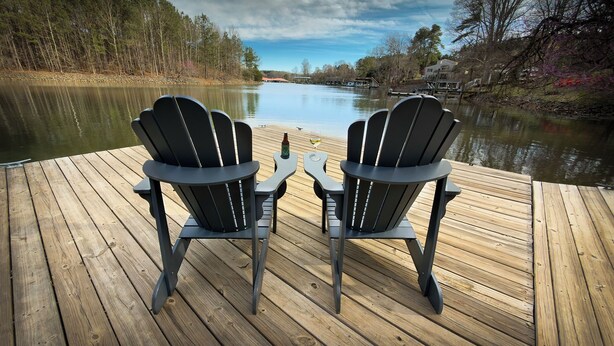 Zwei Adirondack-Stühle auf einer Holzterrasse mit Blick auf einen ruhigen Fluss, im Hintergrund Bäume und ein Haus.