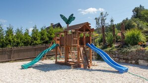 A wooden play structure with two slides in a gravel area, surrounded by greenery and a wooden fence.