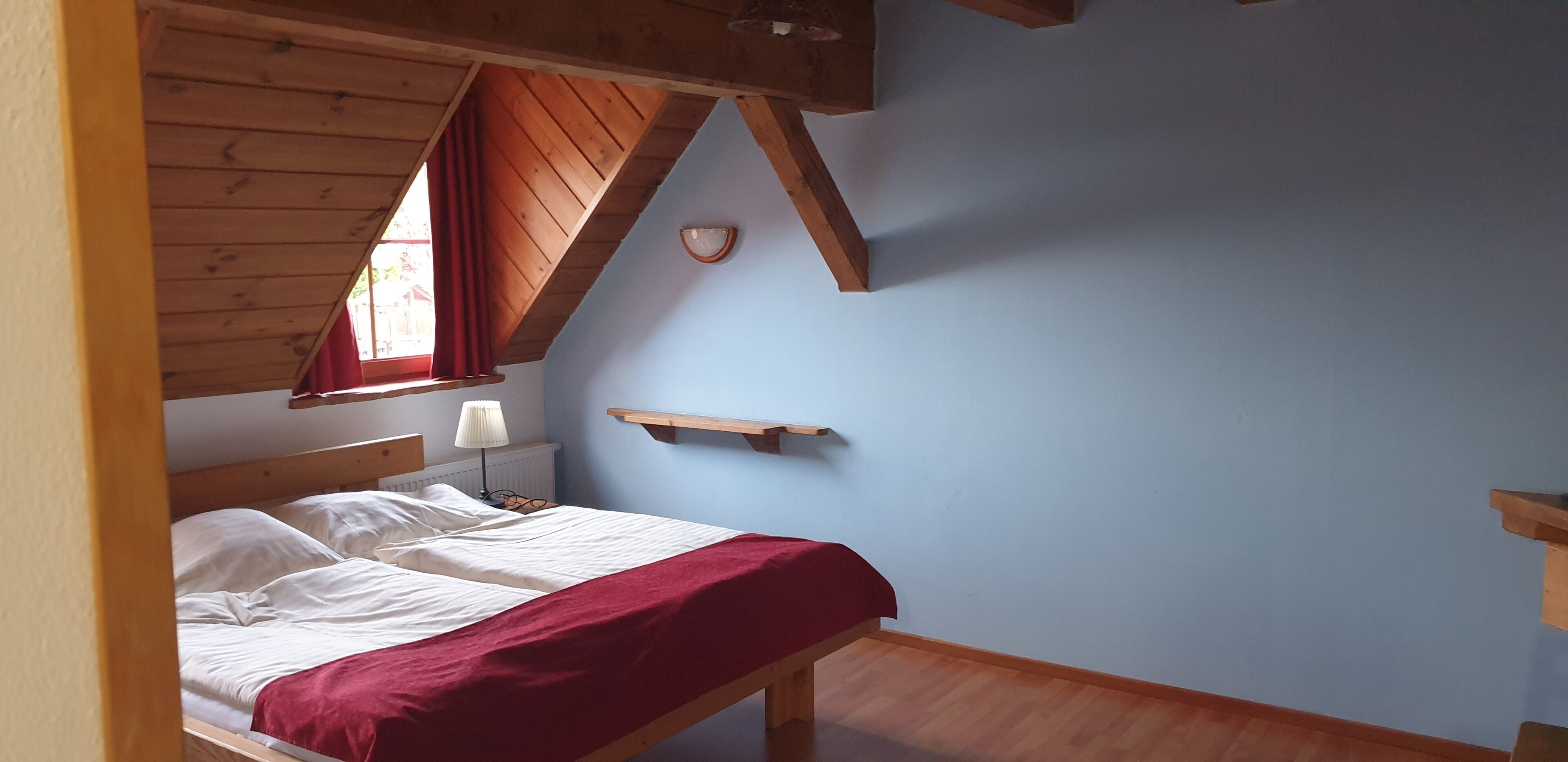 Un dormitorio con una cama de madera, una colcha roja, una lámpara de noche y un estante pequeño.