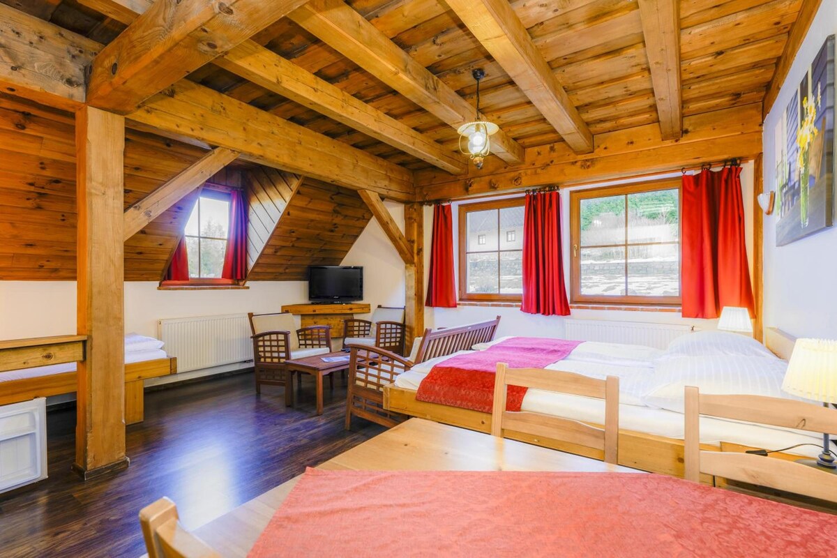 Una habitación acogedora con techo inclinado, vigas de madera, una cama con manta roja, un escritorio con televisor y una ventana con cortinas rojas.
