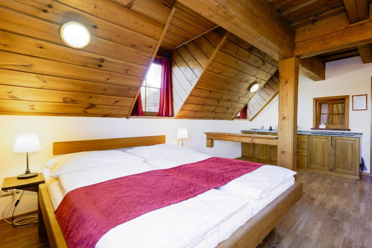 A bedroom with a wooden ceiling, a bed with white bedding and a red blanket, a nightstand with a lamp, and a small window with red curtains.
