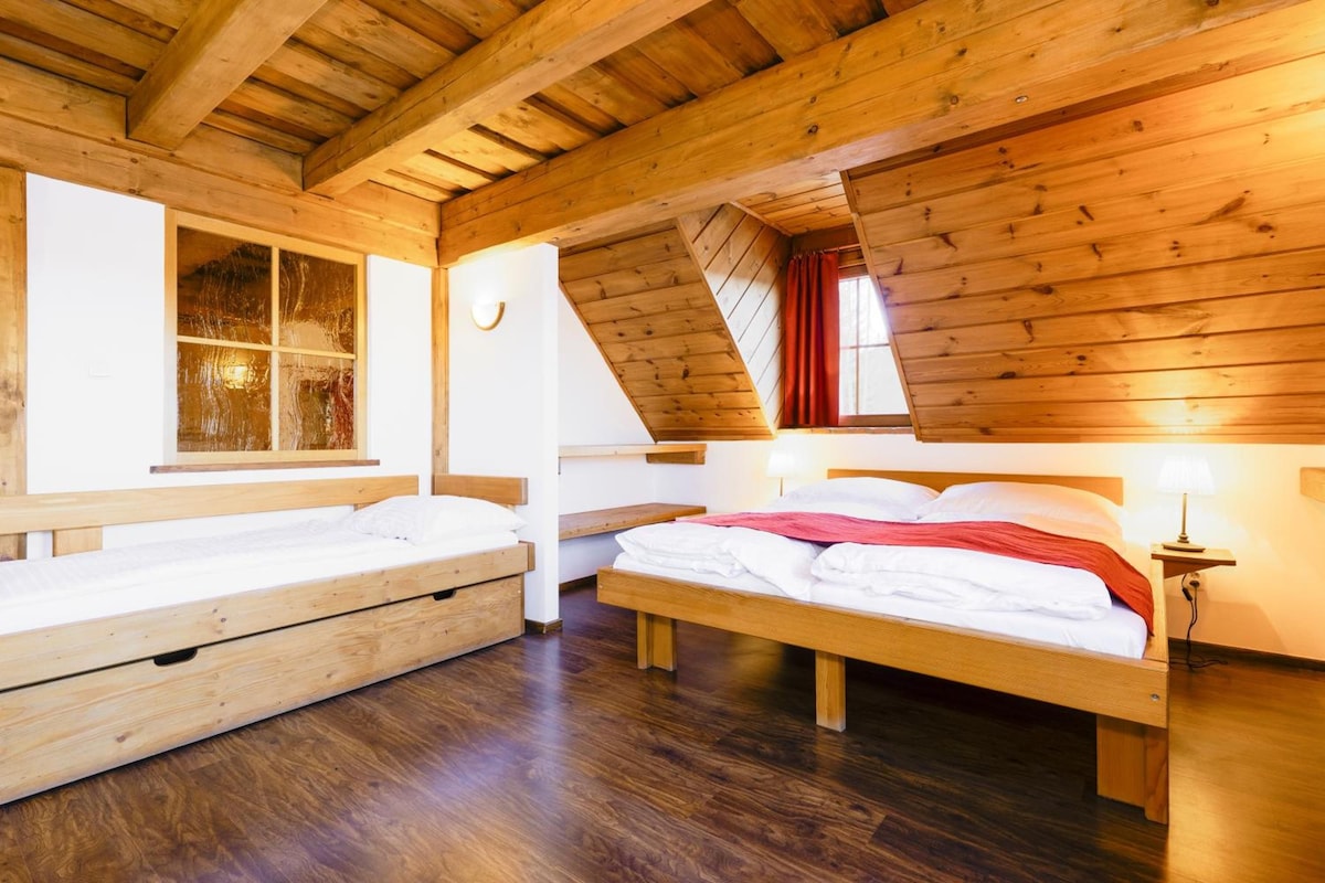 A room with a sloped ceiling, wooden walls, a bed with white bedding, a bench, and a window with curtains.