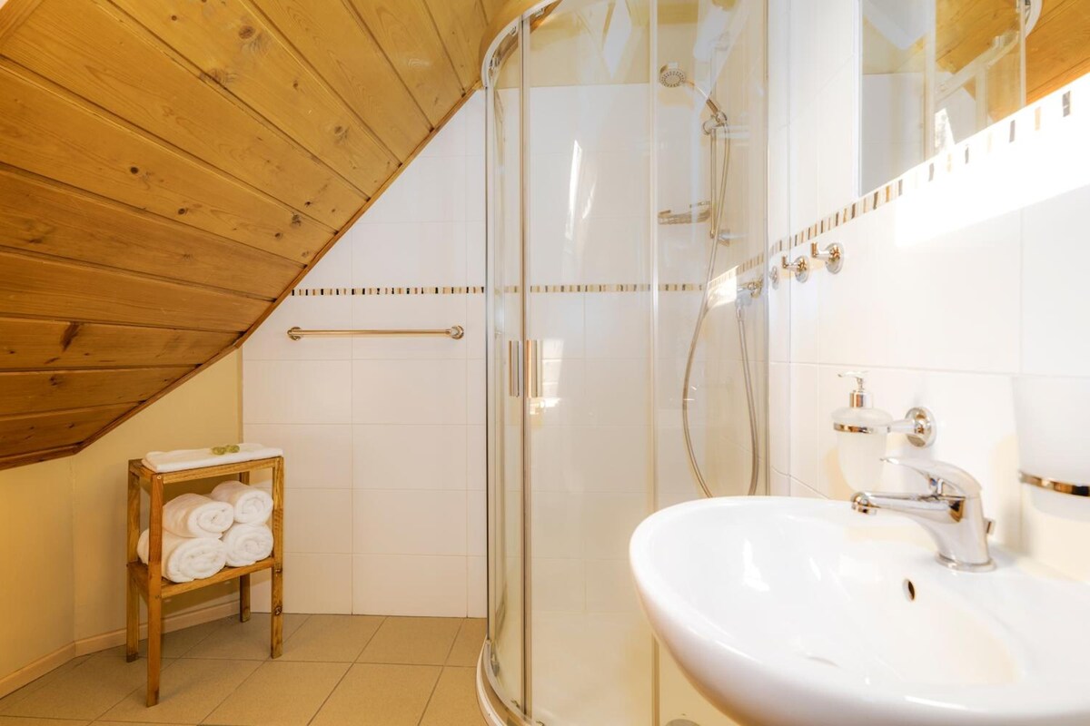 A bathroom with a glass shower enclosure, a wooden bench with towels, and a white sink with a gold faucet.