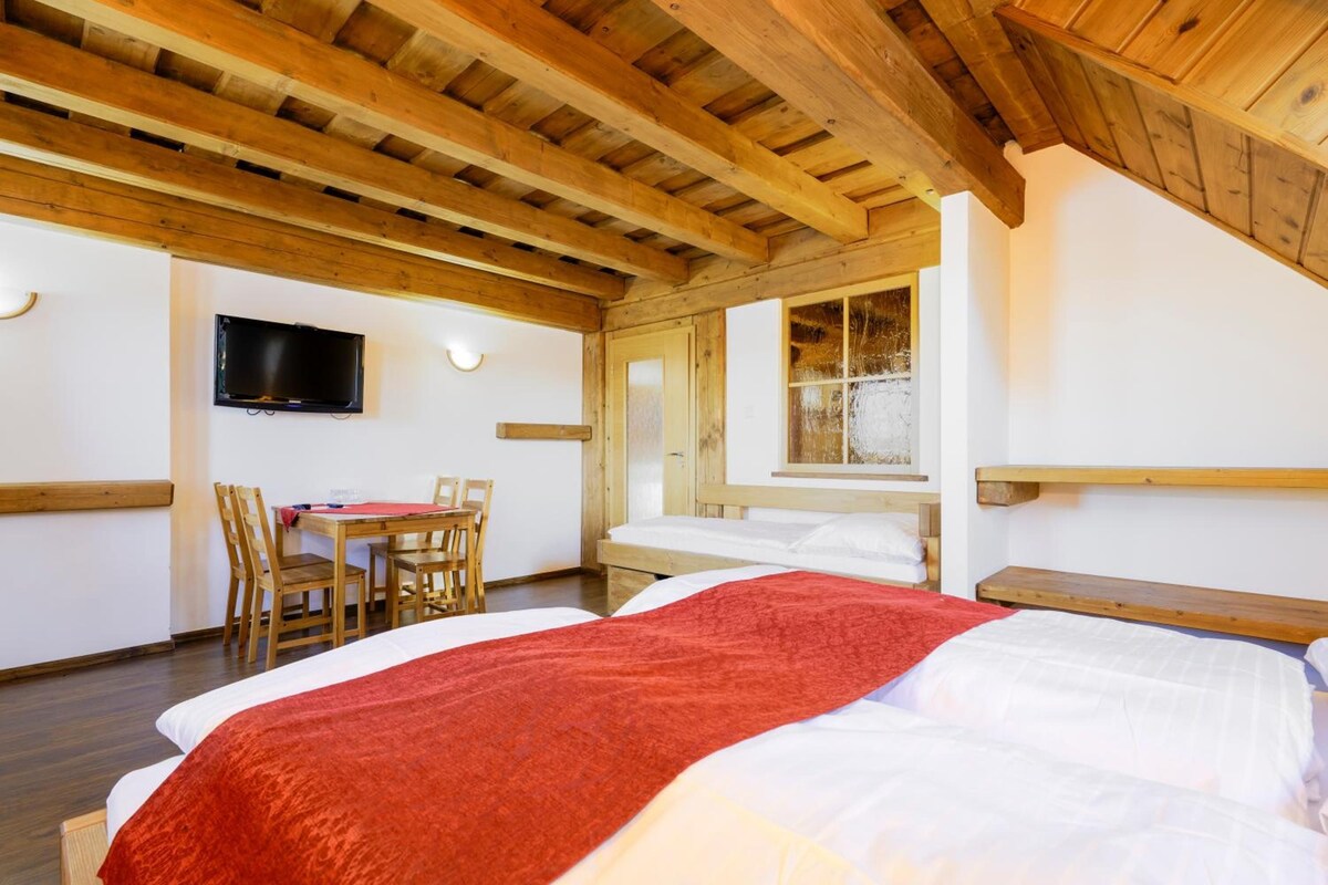 A bedroom with a wooden ceiling, a bed with a red blanket, a TV mounted on the wall, and a window with a view.