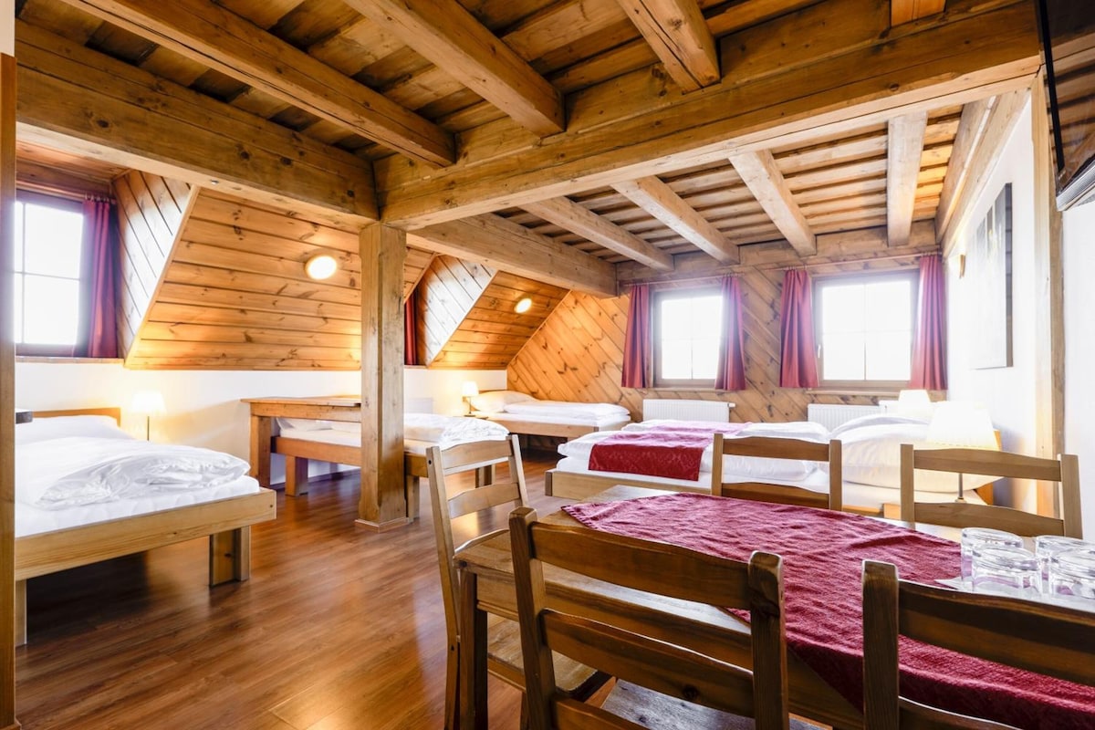 A wooden cabin with four beds arranged in a U-shape, a dining table, and a large window providing natural light.