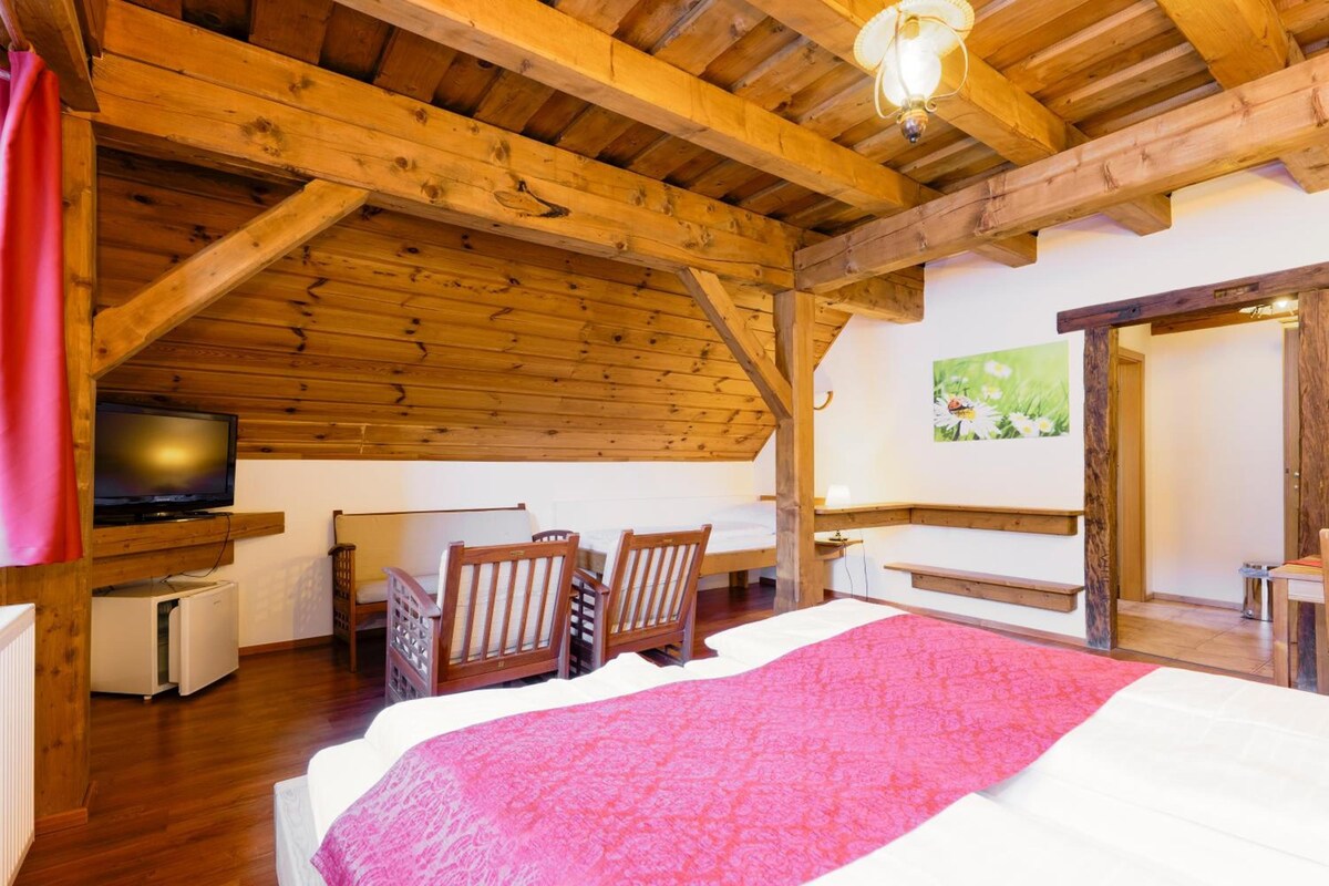 A room with a wooden ceiling, a bed with a pink blanket, a television on a stand, and a painting on the wall.