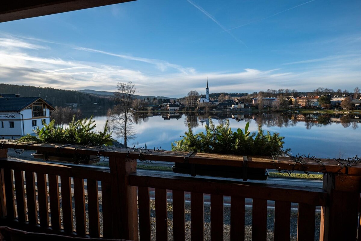 A lakeside view with a wooden deck, a church, and residential buildings in the background.