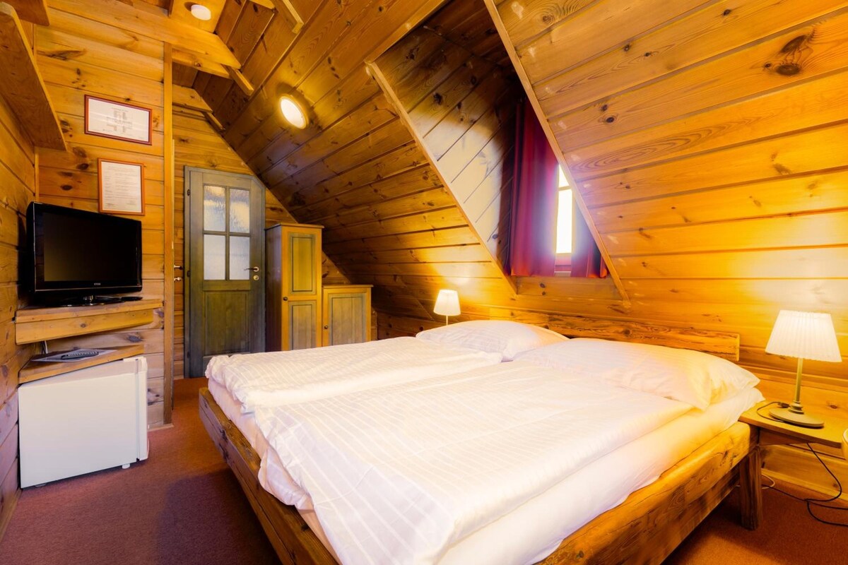 A cozy attic bedroom with a wooden sloped ceiling, a bed with white bedding, a television, and a small refrigerator.