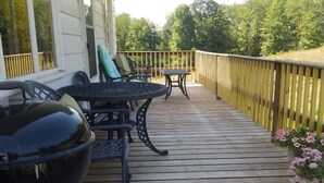 Una terraza de madera con mobiliario exterior, incluyendo una parrilla y sillas, con vista a una zona boscosa.