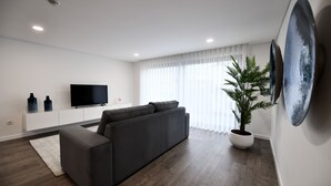 Una sala moderna con un sofá seccional gris oscuro, un televisor de pantalla plana montado en una pared blanca, un helecho en maceta y un ventanal con cortinas traslúcidas.