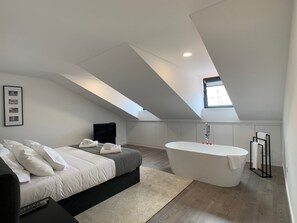 A modern bedroom with a large bed, a flat-screen TV, a skylight, and a freestanding bathtub.
