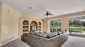 A spacious living room with a large sectional sofa, a ceiling fan, and a view of a swimming pool through large windows. - Family Villa With Pool and Golf, 20 Mins To Disney (Davenport)
