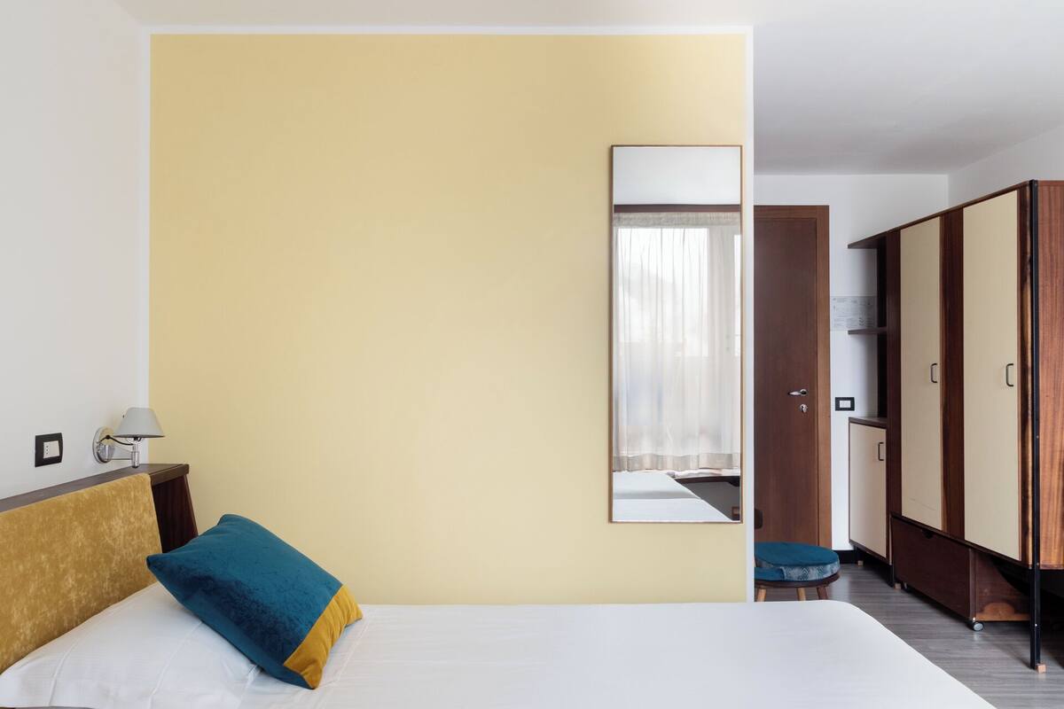 A hotel room with a bed, a nightstand, a lamp, a wardrobe, and a mirror on the wall.