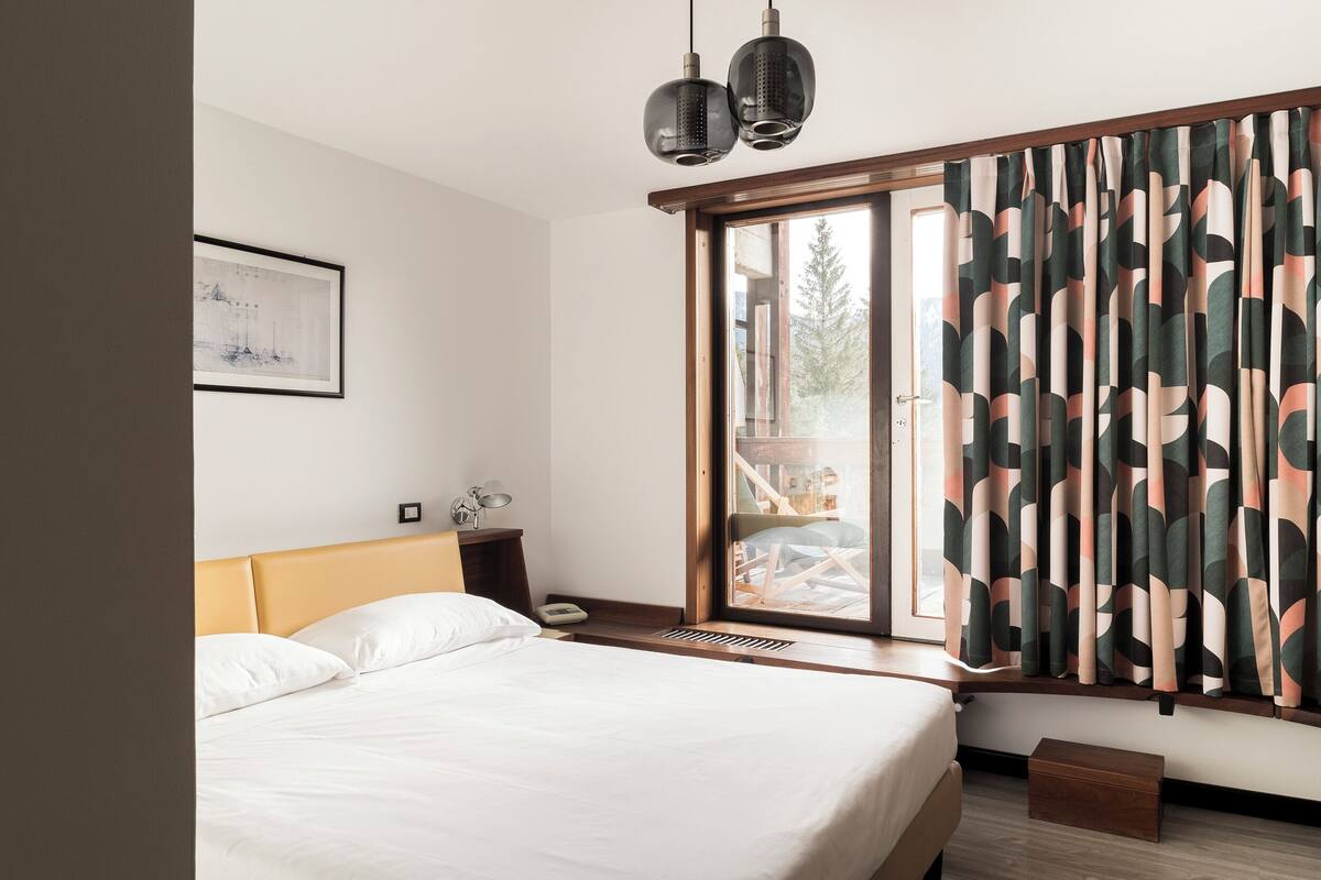 A modern hotel room with a large bed, a bedside table with a lamp, a framed picture on the wall, and a window with curtains.