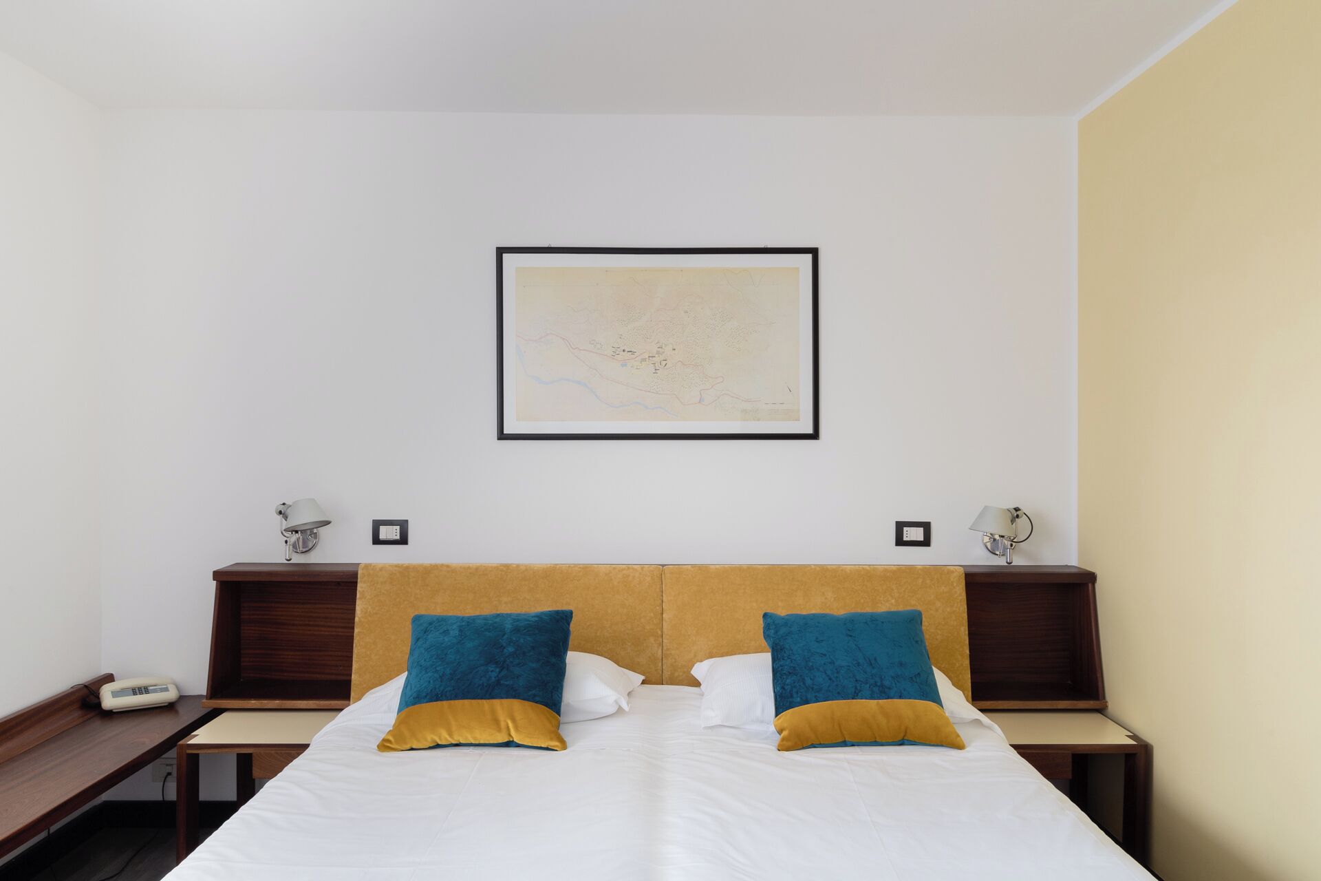 Une chambre d’hôtel comprenant un lit, deux tables de chevet, un tableau abstrait encadré et une lampe fixée au mur.
