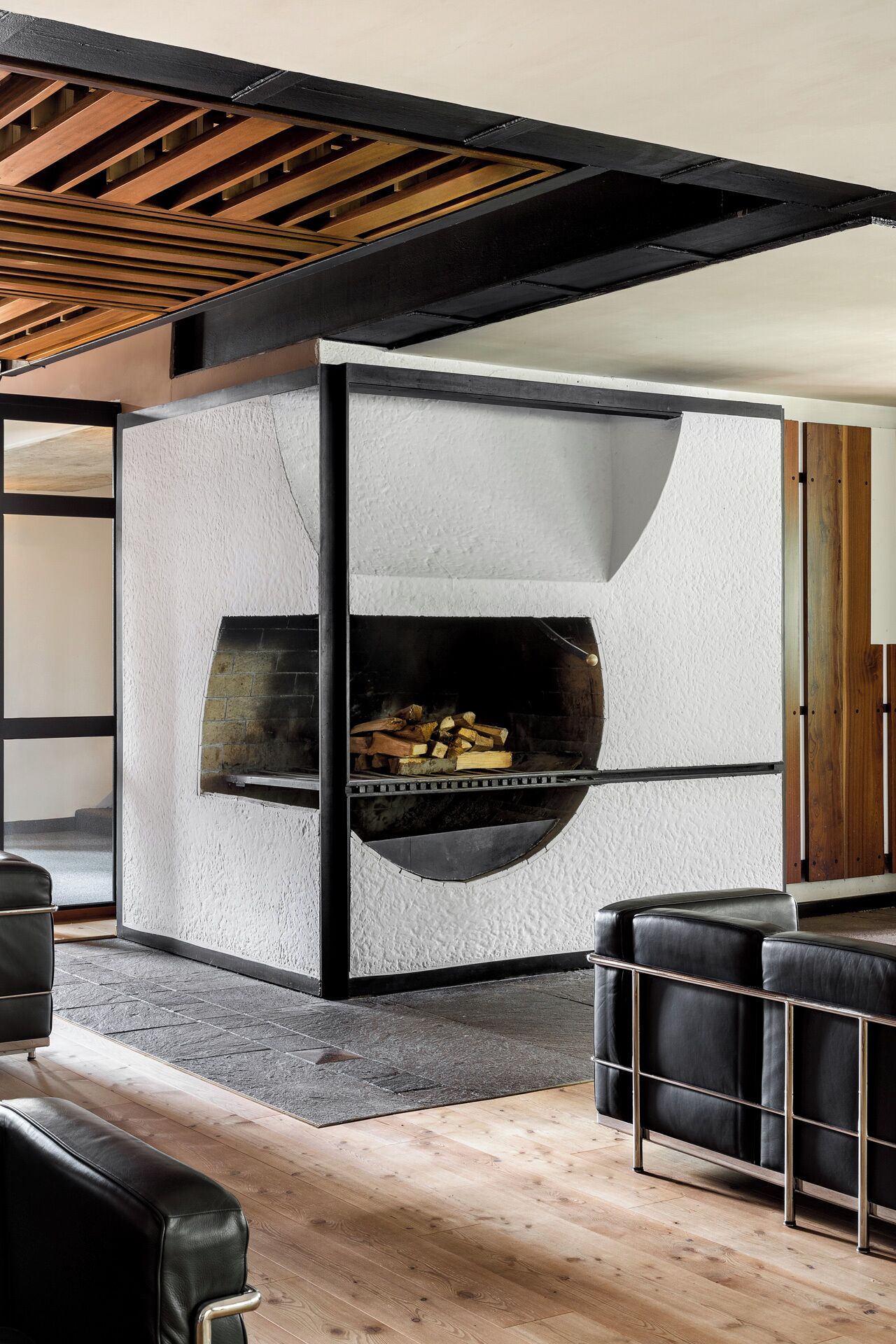 A modern living room with a built-in fireplace, black leather furniture, and a wooden ceiling structure.