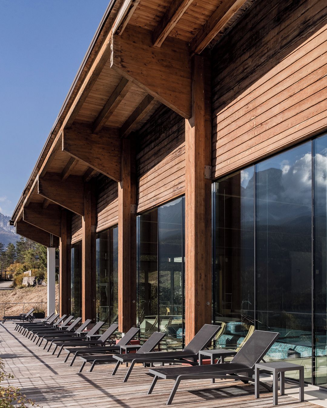 Een houten gebouw met grote glasramen en een terras met loungestoelen.