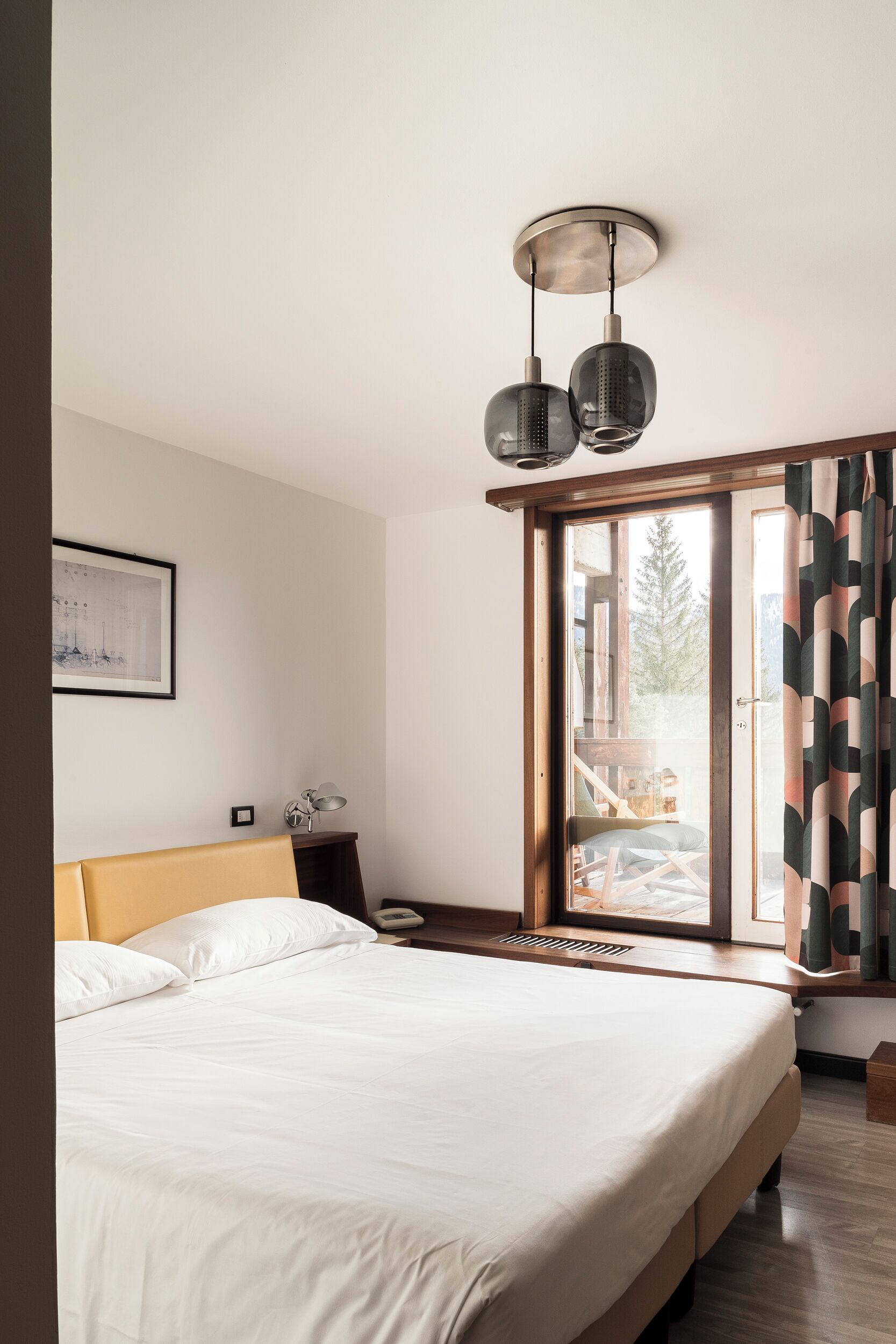 Une chambre à coucher avec un lit, une table de chevet, une lampe, un tableau encadré au mur et une fenêtre avec des rideaux.