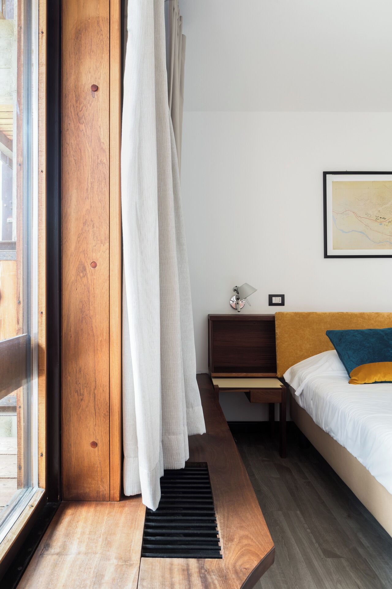 A modern bedroom with a wooden headboard, a bed with white and blue bedding, a nightstand, a wall-mounted lamp, and a window with curtains.