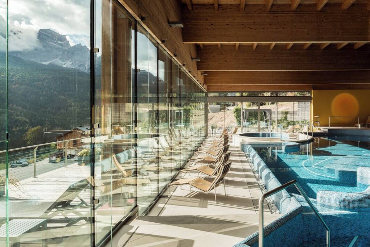 A modern indoor pool area with glass walls, lounge chairs, and a view of mountains.