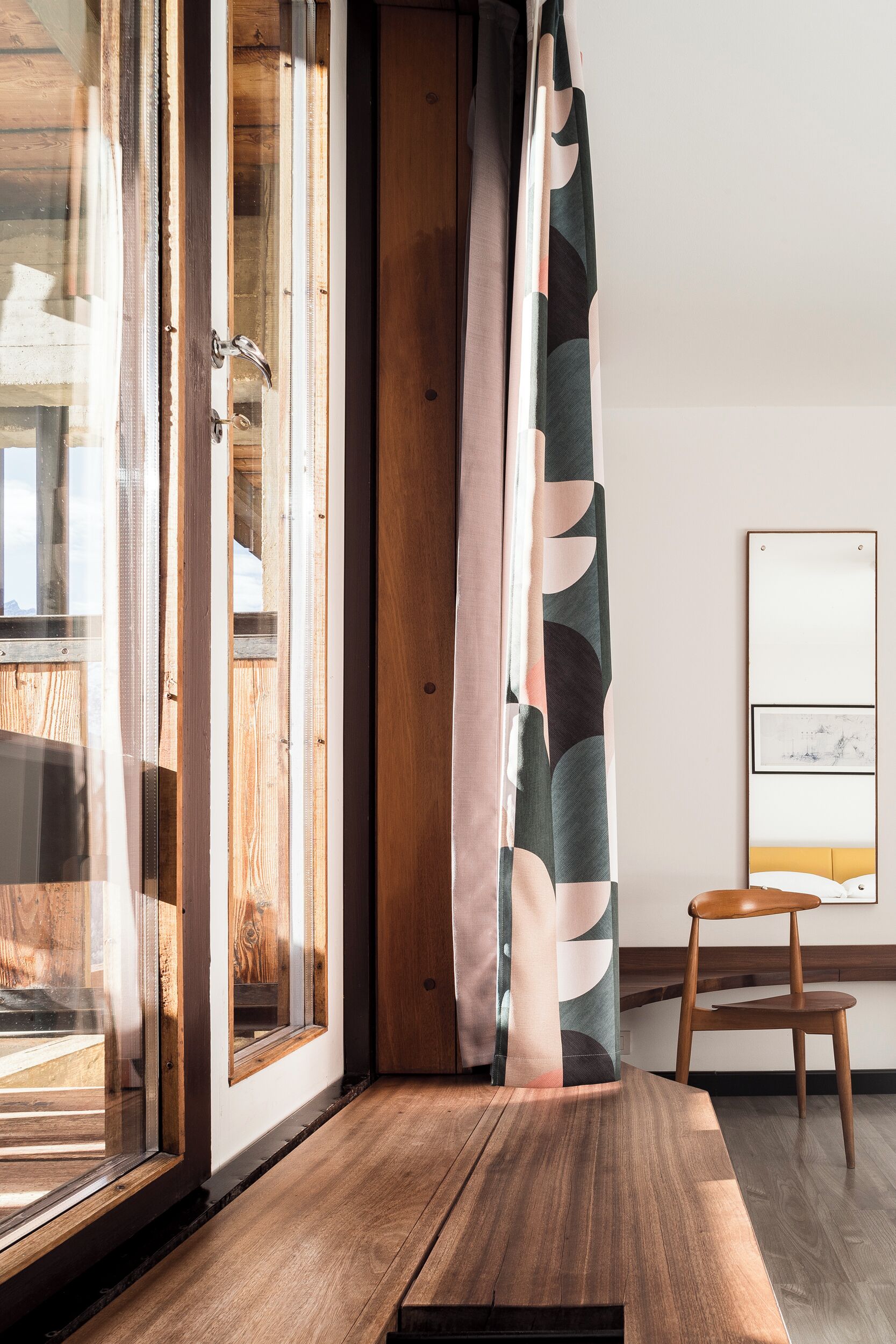 Une pièce avec un plancher en bois, une porte en verre, une chaise en bois et un rideau orné d’un motif de feuilles.