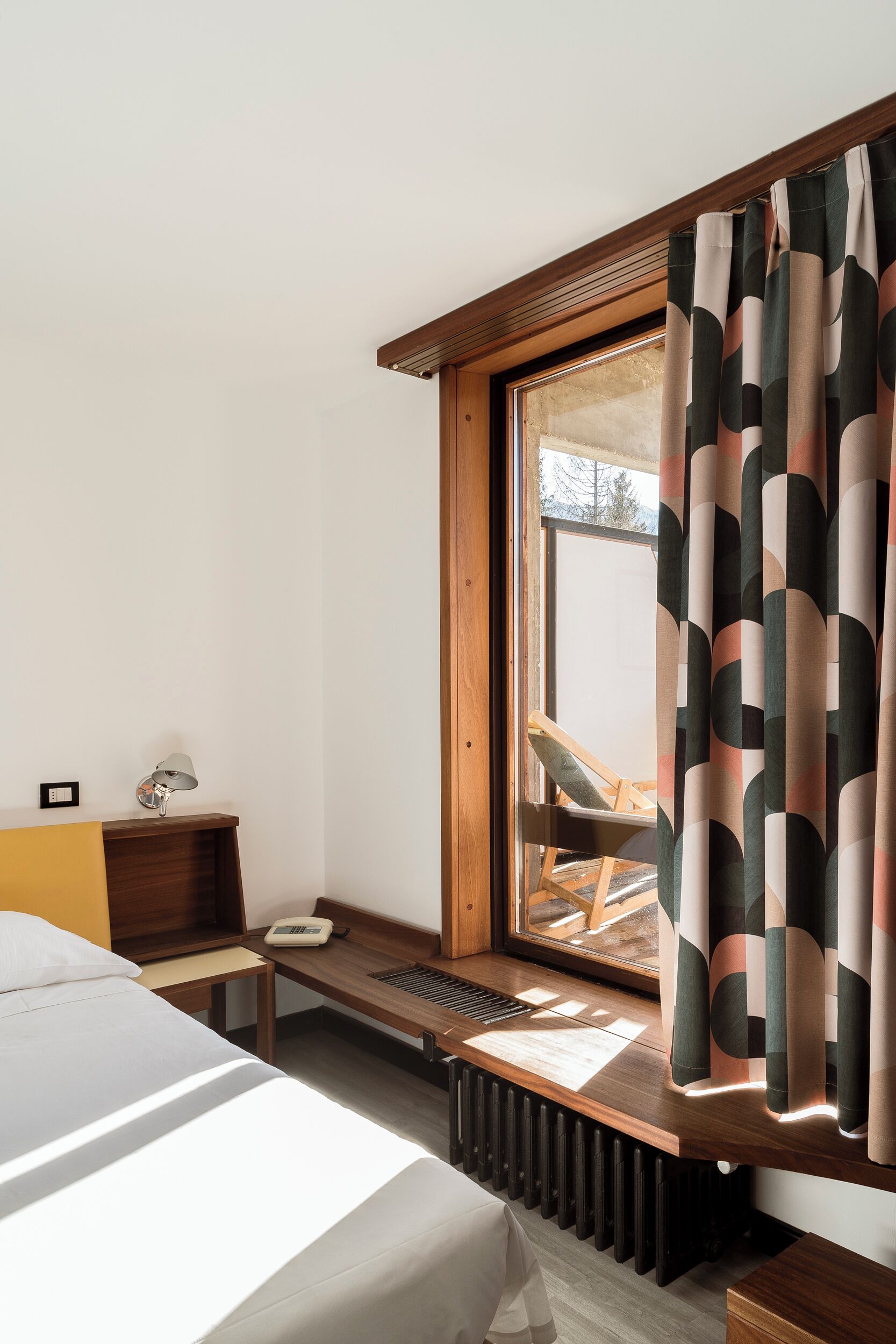 A hotel room with a bed, bedside table, lamp, and a window with a view of an outdoor deck.