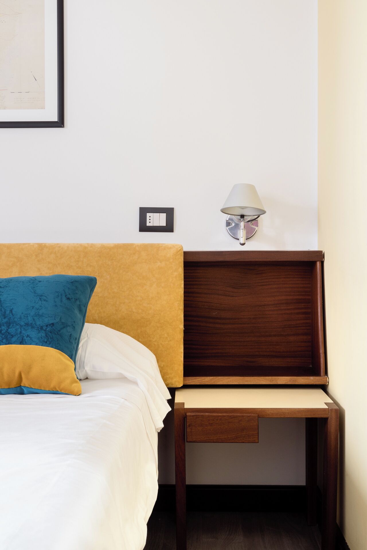 A neatly made bed with a blue and yellow pillow, a wooden nightstand with a lamp, and a wall-mounted light switch.