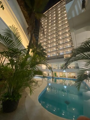 An indoor swimming pool with a curved edge, surrounded by potted plants and palm trees. - Bnow Hotels Acapulco (Acapulco)
